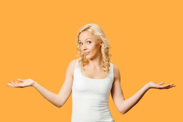 attractive curly blonde woman showing shrug gesture isolated on orange  - Фото, изображение