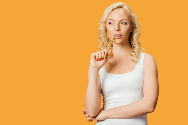pensive blonde woman holding pencil near mouth while thinking isolated on orange  - 写真・画像