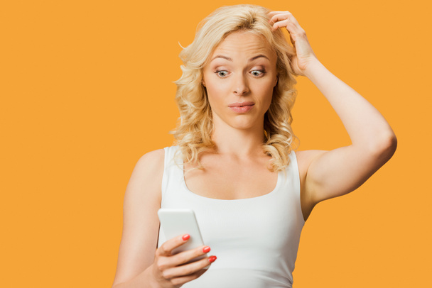 mujer rubia sorprendida mirando el teléfono inteligente y tocando el cabello aislado en naranja
  - Foto, Imagen