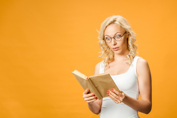 surprised blonde woman in glasses reading book isolated on orange  - Фото, зображення