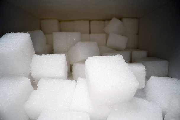 Macro shot of sugar cubes next to carton box on wooden table. Unhealthy food sweetener, sweet crystal cubes - Photo, Image
