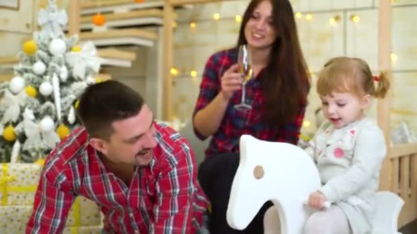 little girl swings on rocking horse beside her parents celebrating Christmas - Footage, Video