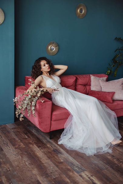 beautiful bride in wedding dress with flowers in hand posing on pink sofa   - Фото, изображение