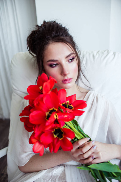 beautiful bride in wedding dress holding bouquet of red tulips and sitting on white sofa  - Fotó, kép