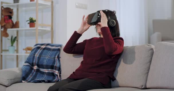 A old woman sitting on the sofa and using a virtual reality glasses to watching something she are very impressed and enthusiastic about this new technology - Filmati, video
