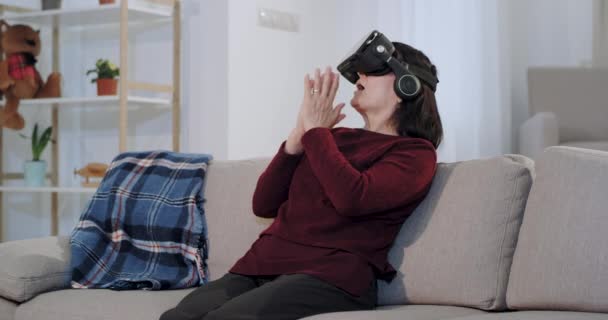 Impressed and enthusiastic old woman using a virtual reality glasses she travel around the world , while sitting on the sofa - Séquence, vidéo