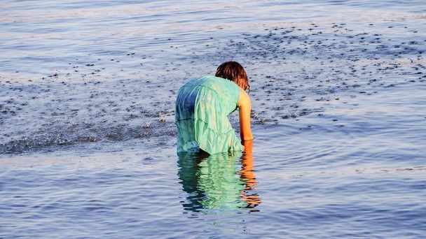   女の子は大西洋でひざまずいて立ち上がり、後ろの景色の中で水の中で手をつないで遊ぶ.  - 写真・画像