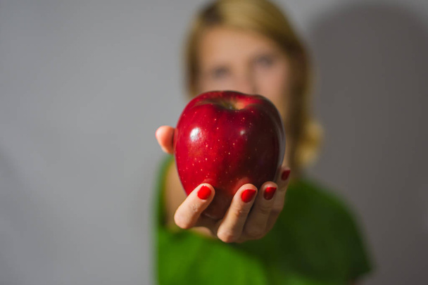 The girl holds a big red apple in hand on a gray background. Apple and fingers in focus. Girl's face and body in defocusing - Φωτογραφία, εικόνα