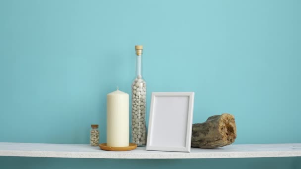Moderna decoración de la habitación con marco de imagen maqueta. Estante blanco contra pared de color turquesa pastel con vela y rocas en botella. Planta de orquídea en maceta
 - Imágenes, Vídeo