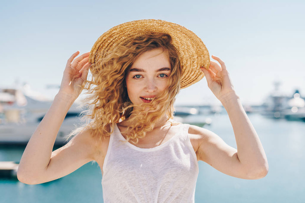 Beautiful young curly woman blonde on the background of the sea and the water holds on to the edges of the hat and laughs - Foto, Imagem