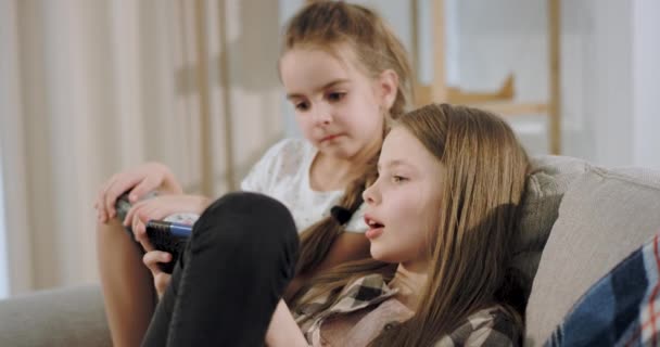 Portrait of a two charismatic girls playing together on a game using a smartphone while sitting on the sofa granny reading a book beside them and looking at her grandchildren. 4k - Felvétel, videó