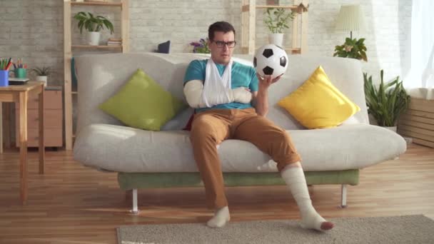 Sad injured athlete sitting on the couch with a soccer ball - Кадри, відео