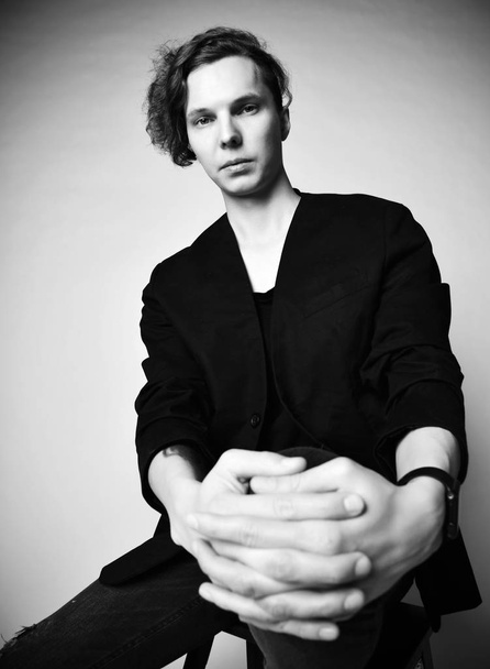 Portrait. Black and white. Young stylish man with creative haircut is sitting on a high chair with hands on his knee  - Фото, зображення