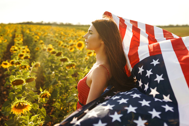 Beautiful girl in hat with the American flag in a sunflower field. 4th of July. Fourth of July. Freedom. Sunset light The girl smiles. Beautiful sunset. Independence Day. Patriotic holiday. - Фото, изображение
