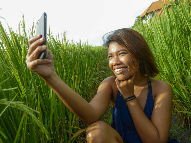 young happy and exotic islander Asian girl from Indonesia taking selfie self portrait photo with mobile phone smiling cheerful and excited posing rice field nature background - 写真・画像