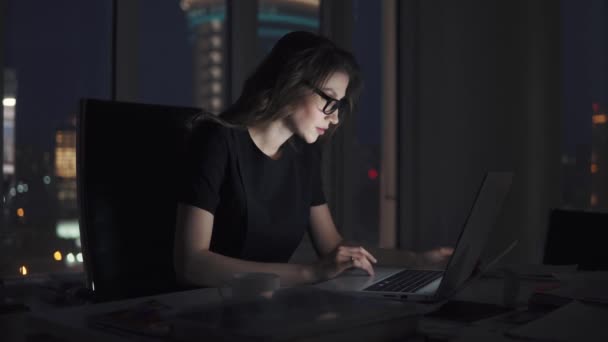 retrato de una joven que trabaja hasta tarde en la oficina. mujer de negocios en gafas trabaja en un ordenador portátil en el fondo de la ciudad de la noche
 - Imágenes, Vídeo