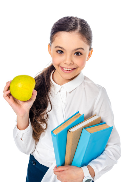 χαμογελαστή μαθήτρια κρατώντας μήλο και βιβλία ενώ κοιτάζοντας την κάμερα απομονωμένη σε λευκό - Φωτογραφία, εικόνα