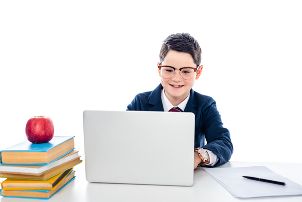 schoolboy in glasses sitting at desk with books and using laptop isolated on white - 写真・画像
