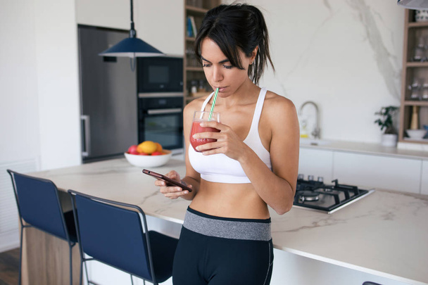 Sporty young woman using her mobile phone while drinking strawberry smoothie in the kitchen at home. - Fotografie, Obrázek