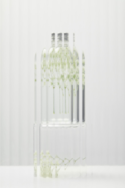 blurred organic cosmetic product in transparent bottle with wildflowers behind glass on grey background - Fotografie, Obrázek