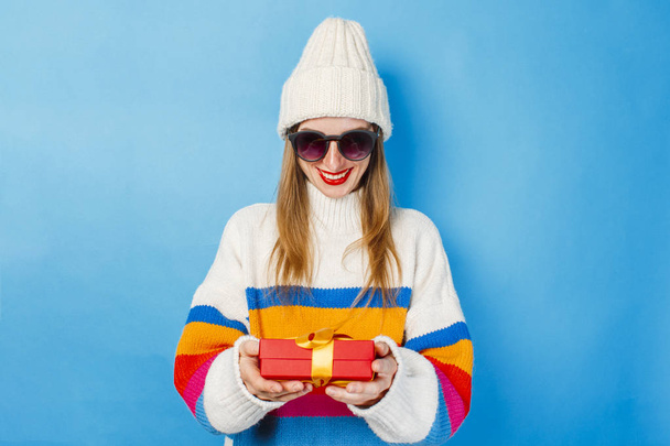 A young girl in glasses, a white hat and a sweater holds in her hands a gift on a blue background. The concept of receiving a gift for the holiday, Christmas, New Year. - Foto, Imagen