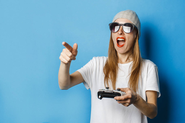 Chica en gafas, sombrero y camiseta blanca sostiene un mando en sus manos y apunta a la pantalla del televisor sobre un fondo azul. Concepto de juego en la consola, deportes cibernéticos, entretenimiento
. - Foto, imagen