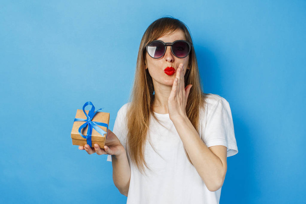 Ένα νεαρό κορίτσι με ένα χαμόγελο στα γυαλιά και ένα λευκό μπλουζάκι κρατά ένα κουτί δώρου σε μπλε φόντο. Έννοια δώρου γενεθλίων ή διακοπών. - Φωτογραφία, εικόνα