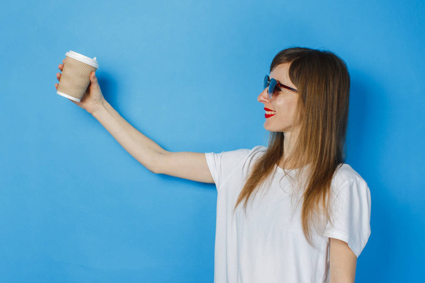 Una joven con gafas y una camiseta blanca sonríe y sostiene una taza de papel con té o café sobre un fondo azul. Concepto cafetería, café para la alegría
. - Foto, Imagen