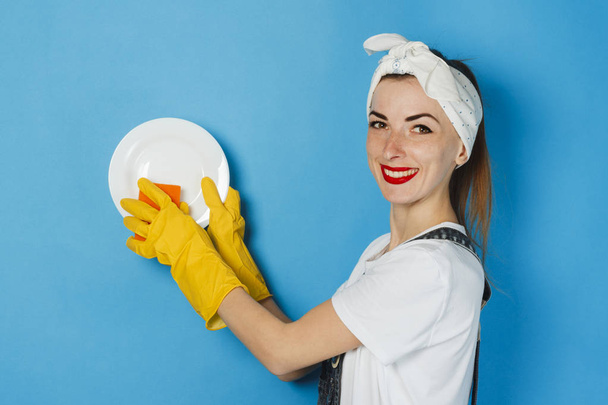 Молодая девушка с шарфом на голове и жёлтыми резиновыми перчатками держит белую тарелку и губку на синем фоне. Концепция уборки и уборки, мытья посуды
. - Фото, изображение