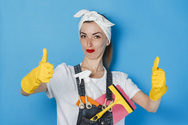 Chica joven con una bufanda en la cabeza y guantes de goma amarillos mostrando un gesto de pulgares hacia arriba, super, como en un fondo azul. Concepto de servicio de limpieza y limpieza, alta calidad
. - Foto, Imagen