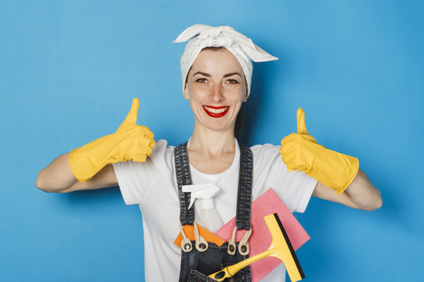 Jong meisje met een sjaal op haar hoofd, gele rubberen handschoenen en schoonmaakspullen met een duim omhoog gebaar, super, als op een blauwe achtergrond. Concept van reiniging en reiniging, hoge kwaliteit. - Foto, afbeelding