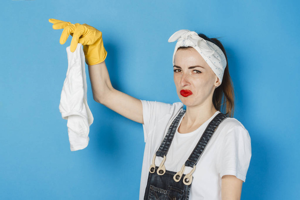 A young girl with a scarf on her head, yellow rubber gloves holds in her hand men's white panties on a blue background. The concept of cleaning and cleaning service, high quality. - Photo, Image