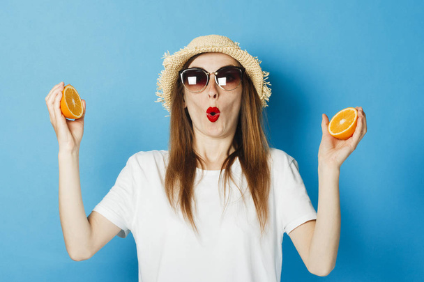 Een jong meisje met een verrast gezicht in glazen en een stro hoed houdt twee halve sinaasappels in haar handen, een blauwe achtergrond. Concept vakantie, toeristische reis, zomer en tropen. - Foto, afbeelding