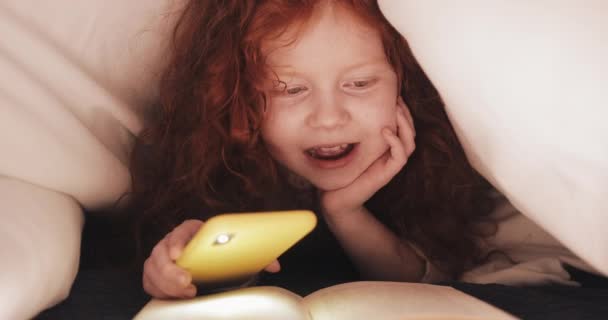 Little redhead girl reading book with interest laying in his bed at home. Girl using flashlight of smartphone for lighting pages. - Séquence, vidéo