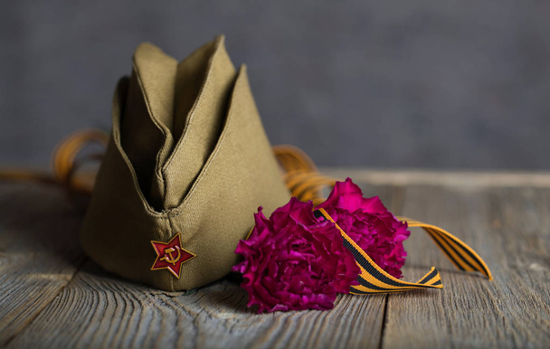 Boné militar, cravos, fita de São Jorge em uma sobremesa de madeira
 - Foto, Imagem