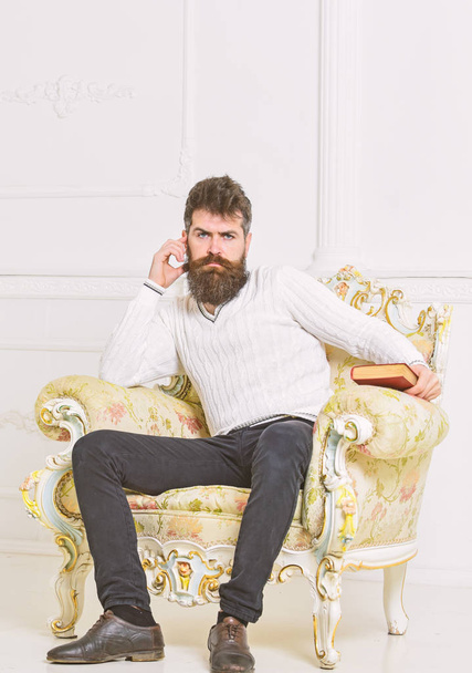 Reflections on literature concept. Connoisseur on thoughtful face finished reading book. Man with beard and mustache sits on armchair, holds book, white wall background. Guy thinking about literature - Фото, изображение