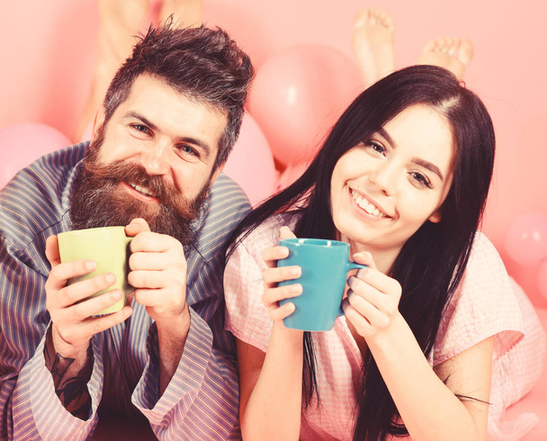 Couple in love drink coffee in bed. Couple relax in morning with coffee. Family tradition concept. Man and woman in domestic clothes, pajamas. Man and woman on smiling faces lay, pink background - 写真・画像