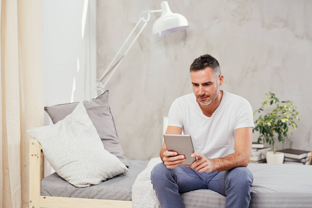 Caucasian man in forties sitting on bed in bedroom and using tablet. - Fotó, kép