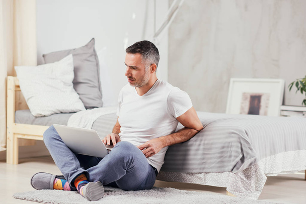 Handsome middle aged man sitting on the floor wirh crossed legs, leaning on bed and using laptop. - Photo, Image