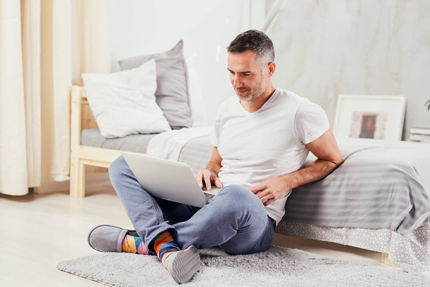 Handsome middle aged man sitting on the floor wirh crossed legs, leaning on bed and using laptop. - Photo, Image