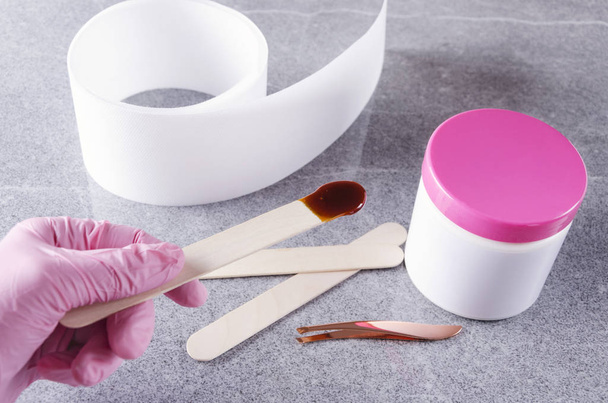ピンクの保護手袋の美容師は、脱毛のためのワックスでスティックを保持します。ワックス処理の調製の概念 - 写真・画像