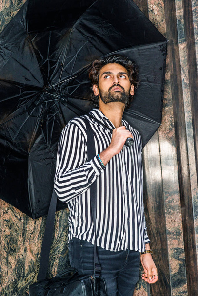 Raining day - grainy, drizzling, wet feel. Young East Indian American Man with beard, wearing black, white striped shirt, carrying bag, holding umbrella, standing outside in New York, looking up - Foto, immagini