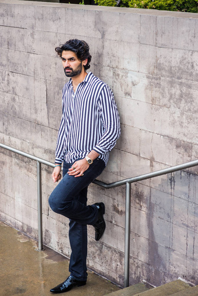 Raining day - grainy, drizzling, wet feel. Young East Indian American Man with beard, wearing black, white striped shirt, black pants, leather shoes, standing against wall outside in New York, thinks - Photo, Image