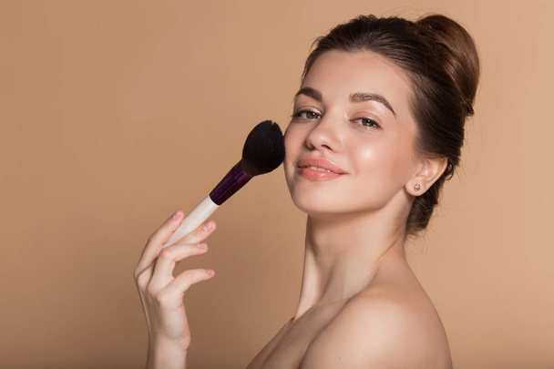Closeup portrait of beautiful happy young girl with perfect skin is holding makeup brush in hand. Isolated on beige. Beauty and cosmetics concept. - Foto, immagini