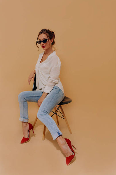 Full-Length portrait of stylish young lady wearing heels and sunglasses sitting over isolated beige background  - Φωτογραφία, εικόνα