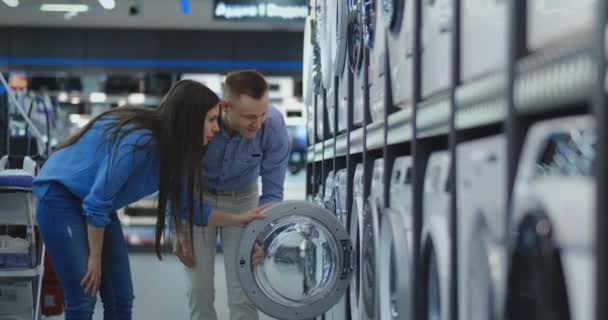 Giovane coppia sposata uomo e donna in negozio di abbigliamento casual elettrodomestici scelgono di acquistare lavatrice per la casa. Aprire la porta guardando nel tamburo, confrontare il design e le caratteristiche dei dispositivi
 - Filmati, video
