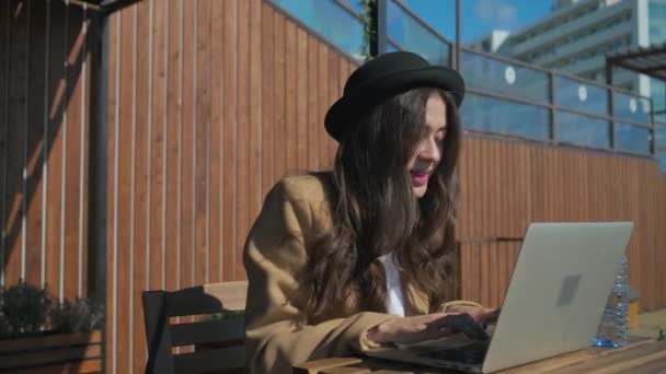 Joyful brunette girl is typing on keyboard of notebook, sitting outdoors - Footage, Video