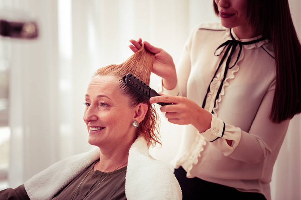 Cheerful hairdresser combing hair of her client - Фото, изображение