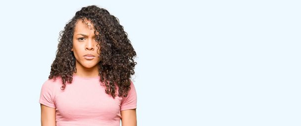 Young beautiful woman with curly hair wearing pink t-shirt skeptic and nervous, frowning upset because of problem. Negative person. - Foto, Imagen