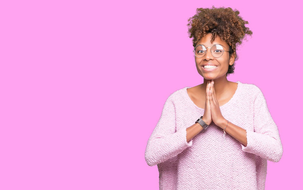 Красивая молодая африканская американка в очках на изолированном фоне молится руками, прося прощения, уверенно улыбаясь
. - Фото, изображение